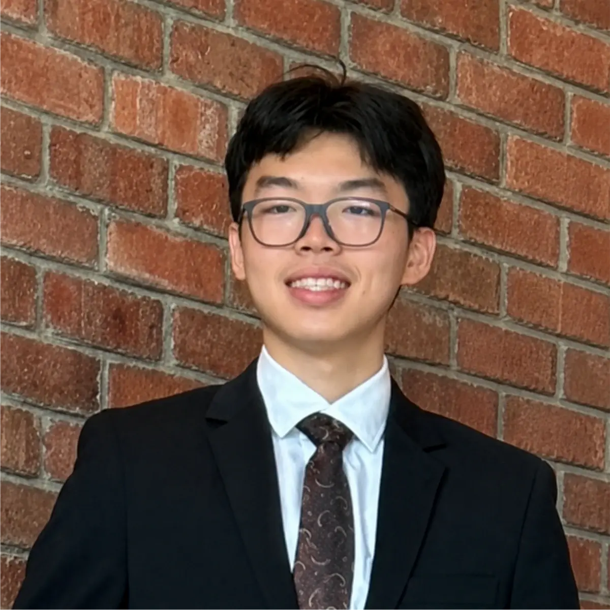 Justin Yu wearing glasses, a dark suit and a patterned tie, against a brick wall.