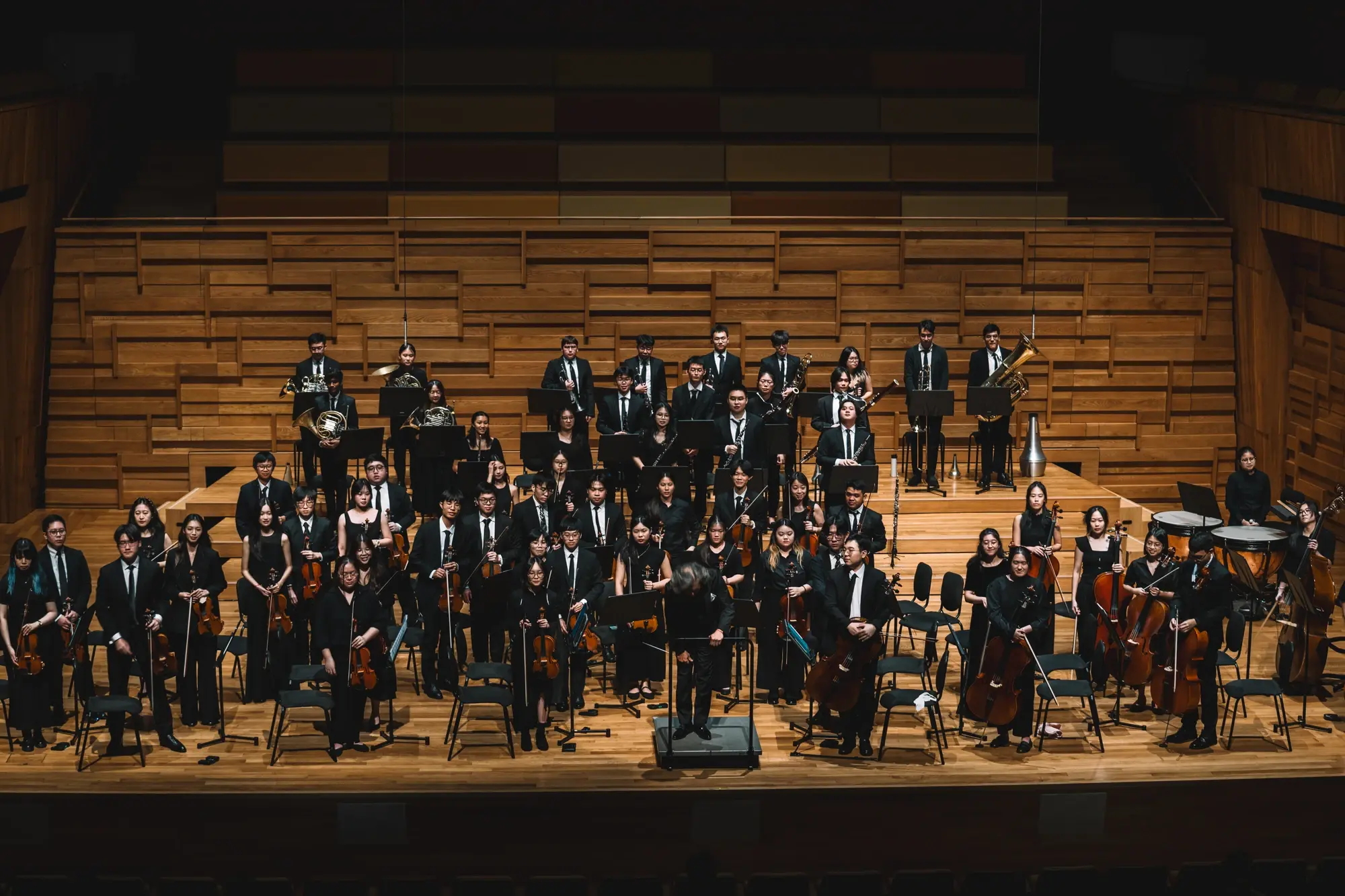 NTU Symphony Orchestra musicians standing with their instruments on a wooden concert stage, with the conductor at the centre.
